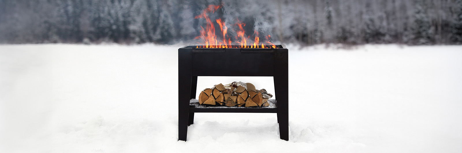 A large outdoor grill for barbequing food. It has fire blazing, with a pile of wood underneath. Snow has settled on the ground and surrounding area. 